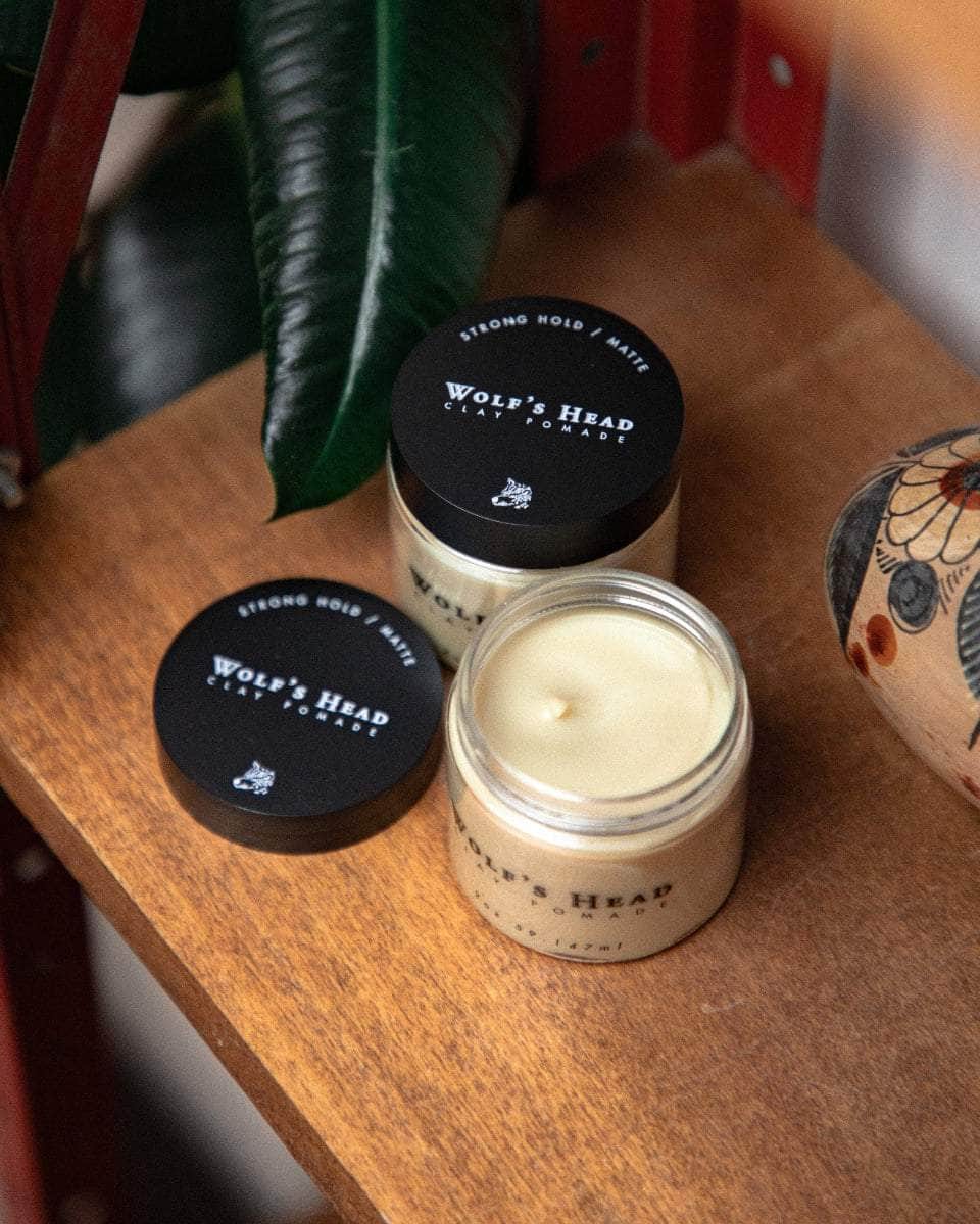 Wolf’s Head Clay Pomade jars on vintage red retail shelf  with plant– strong hold matte finish hair product. Open jar shoes smooth clay in jar.