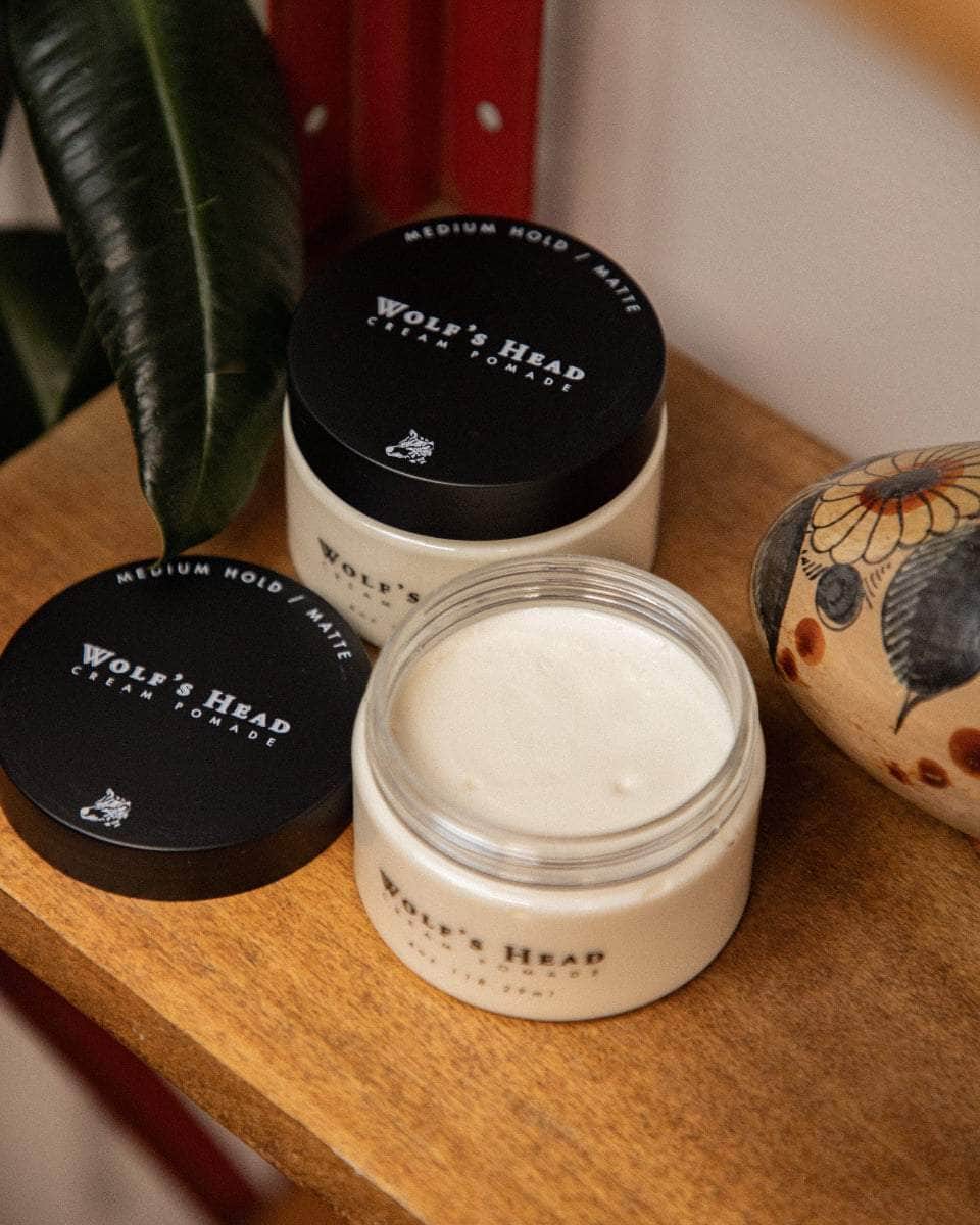 Wolf’s Head Cream Pomade jars on vintage red retail shelf  with plant– medium hold matte finish hair product. Open jar shoes smooth cream in jar.