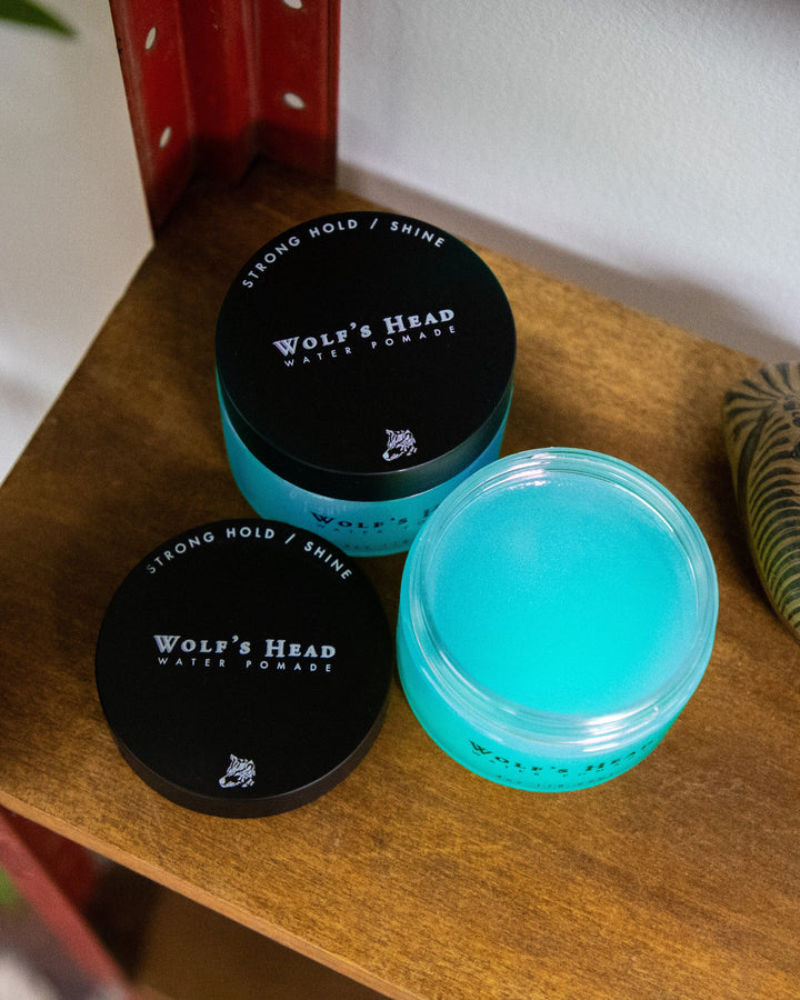 Two containers of Wolf’s Head Water Soluble Pomade on a vintage shelf, one with the lid off and placed beside the jar. Strong hold, high shine, and clean ingredients for premium men’s grooming.